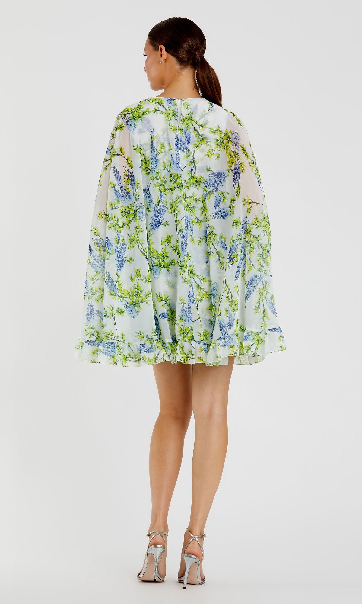 A woman faces away, wearing the Mac Duggal Cape Short Party Dress 56029—a loose, short white floral dress with green and blue patterns—and silver high-heeled sandals against a plain white background.